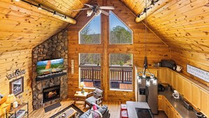 A cabin interior with a stone fireplace, a television mounted on the wall, a dining area, and a kitchen with wooden cabinets and a countertop. - LOG CABIN★SECLUDED AUGUST SPECIALS★HOT TUB★GAME ROOM★CONVENIENT★FIRE PIT (Sevierville)