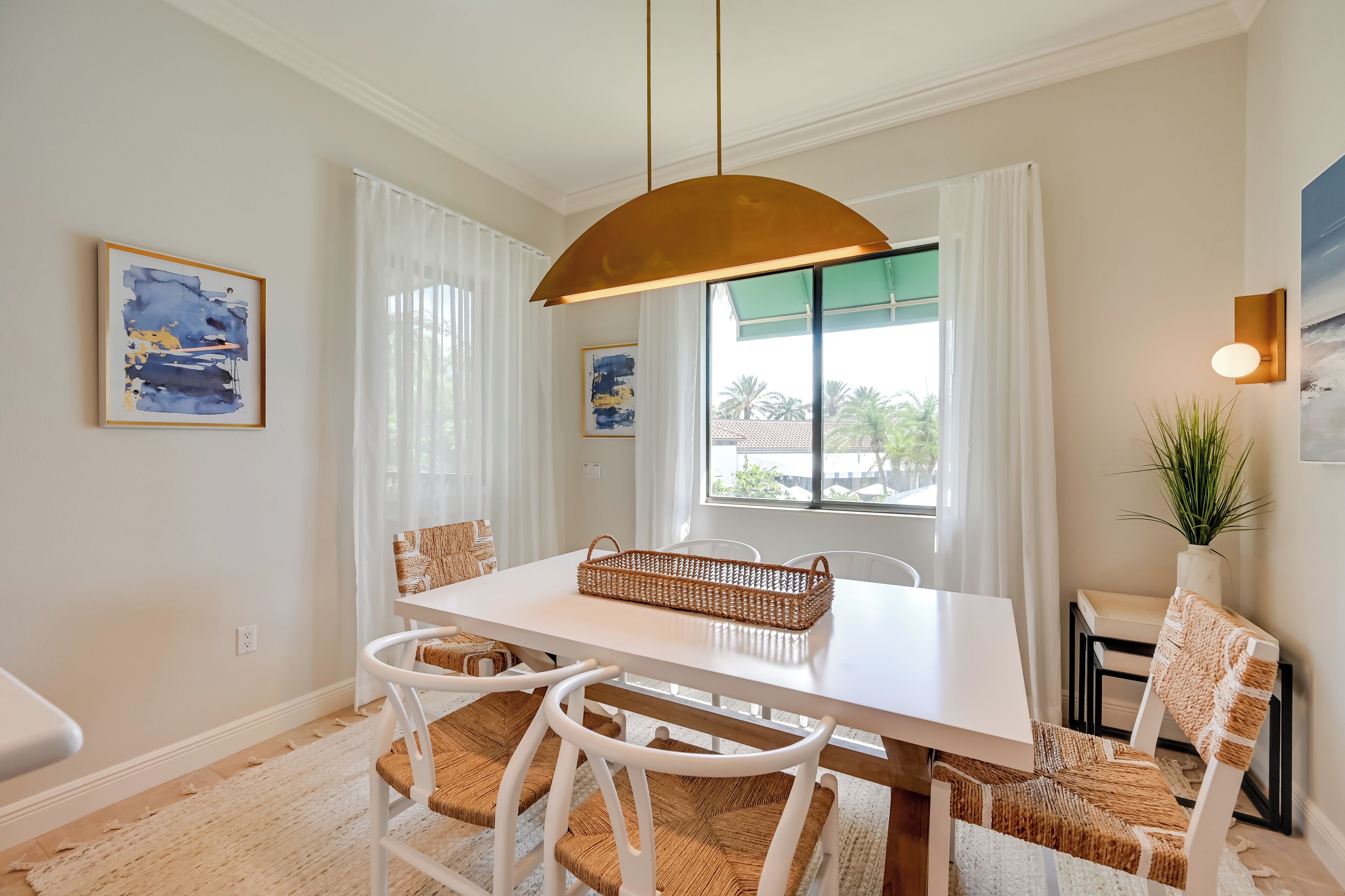 A dining area with a white table, wicker chairs, a hanging light fixture, and artwork on the walls.