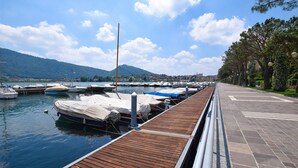 Eine Holzsteg mit anlegenden Booten in einer Marina, ein gepflasterter Weg und eine bergige Landschaft im Hintergrund.