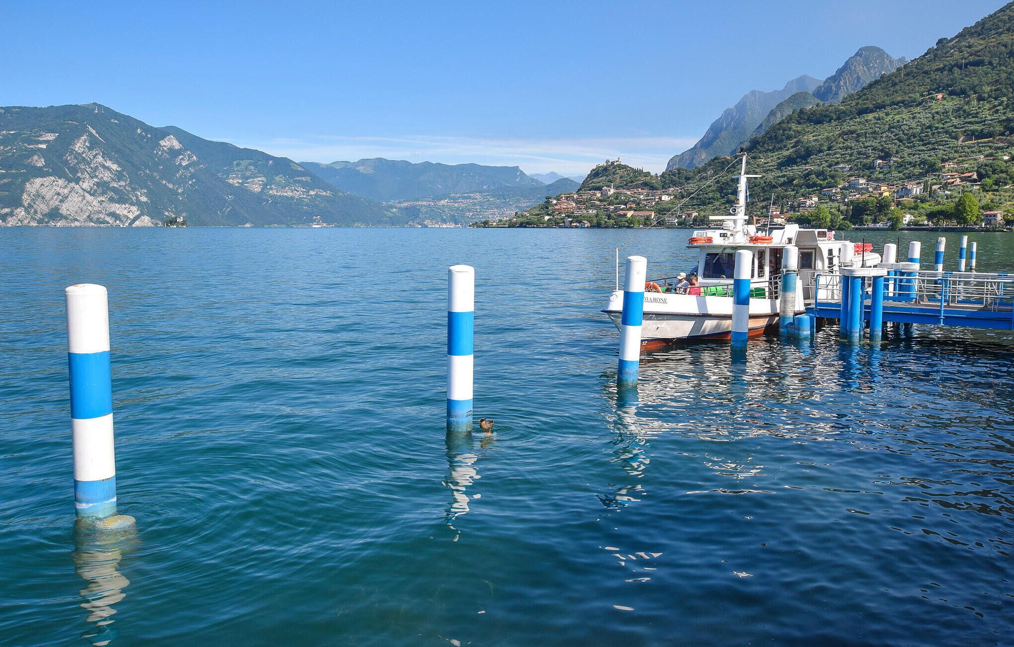 Un barco amarrado en un muelle con topillos azules y blancos, rodeado por un paisaje montañoso y un pueblo al fondo.