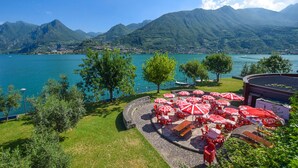 Eine Seeterrasse mit roten und weißen Sonnenschirmen, Steinboden und Blick auf einen See und Berge.