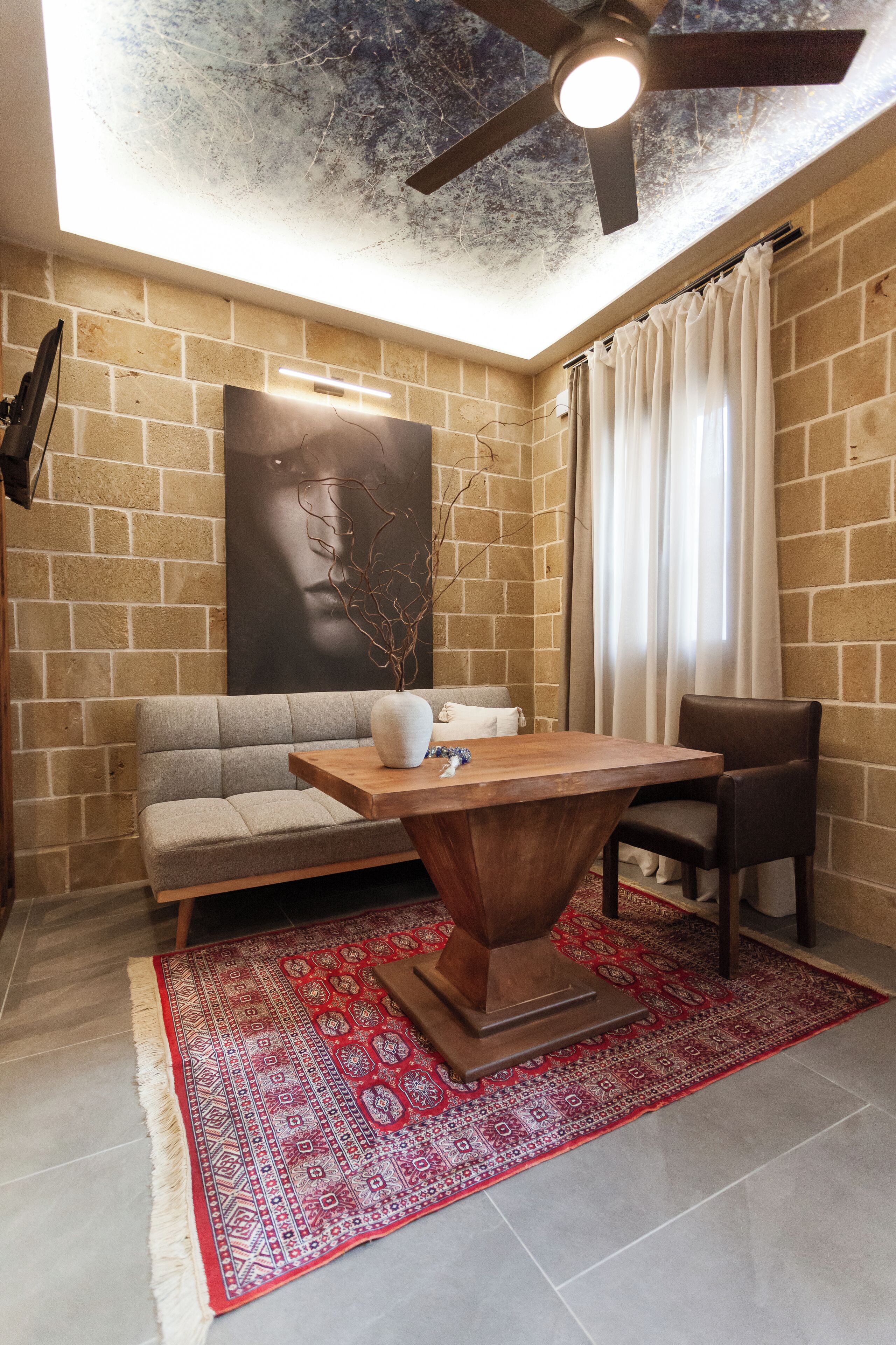 Ein Zimmer mit Backsteinwand, Deckenventilator, Sofa, Tisch, Stuhl und einem gemusterten Teppich.