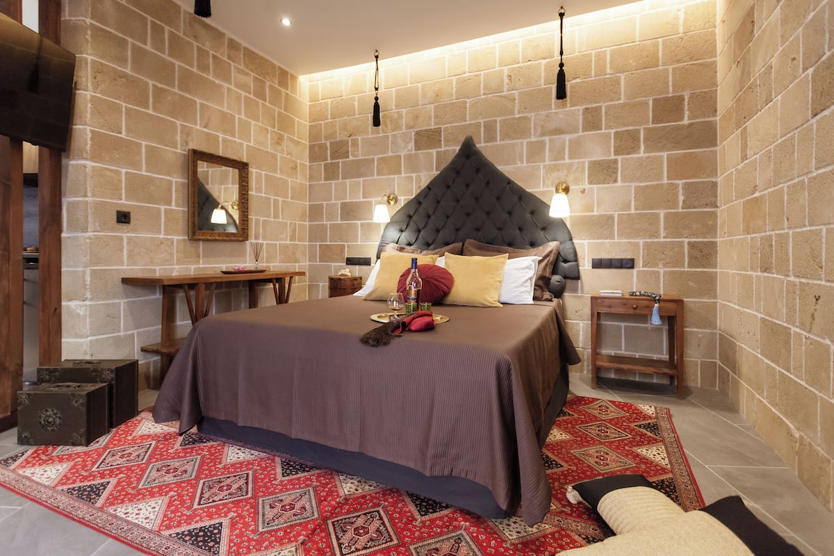 Un dormitorio con una cama grande, una mesita de noche de madera, un espejo y una alfombra roja estampada.