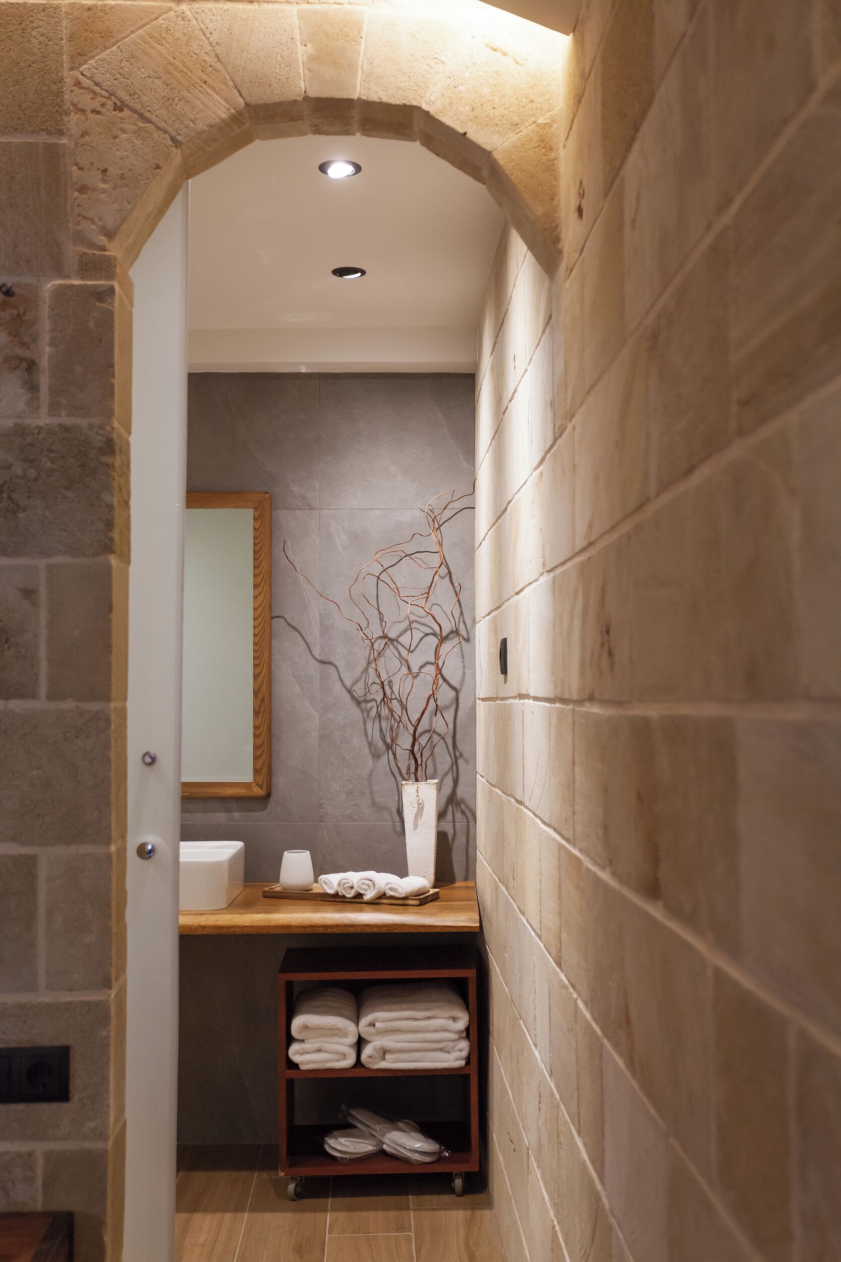 A bathroom with a stone archway, a mirror, a vase with branches, and a shelf with towels.