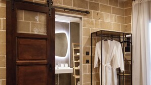 Un baño con puerta de madera, un perchero con batas blancas, un espejo y una botella sobre una bandeja.