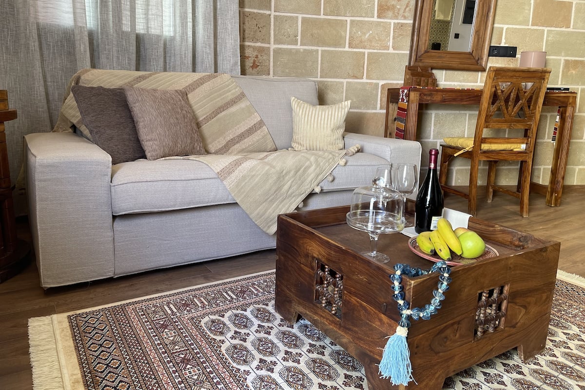 Un salón con un sofá gris, una mesa de centro de madera con frutas y una botella de vino, y una alfombra estampada.