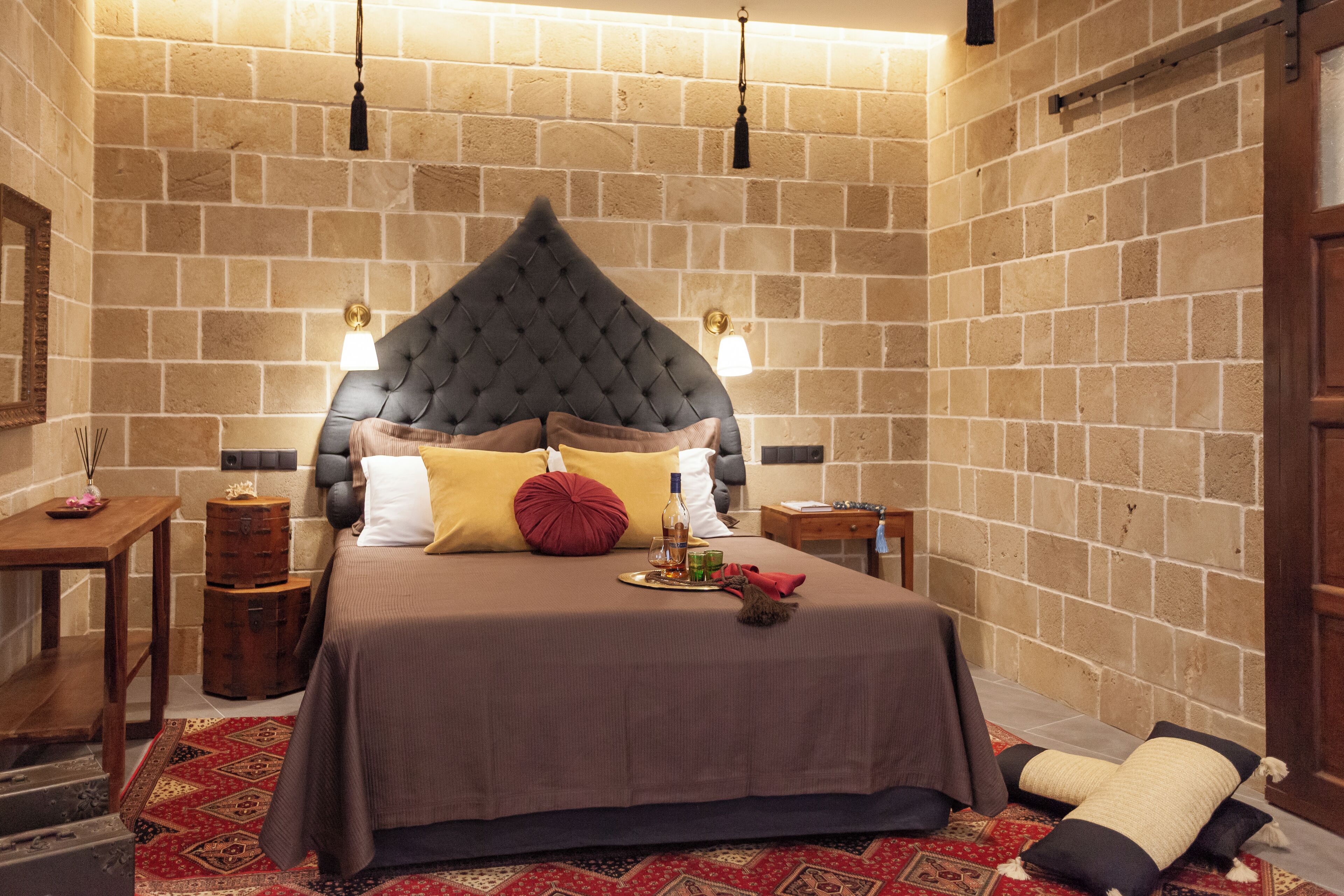 Ein Schlafzimmer mit Backsteinwand, einem gepolsterten Kopfteil, einem Holznachttisch, einem Beistelltisch, einem roten Teppich mit Muster und einem großen Bett mit brauner Bettdecke und Kissen.