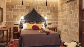 Ein Schlafzimmer mit Backsteinwand, einem gepolsterten Kopfteil, einem Holznachttisch, einem Beistelltisch, einem roten Teppich mit Muster und einem großen Bett mit brauner Bettdecke und Kissen.