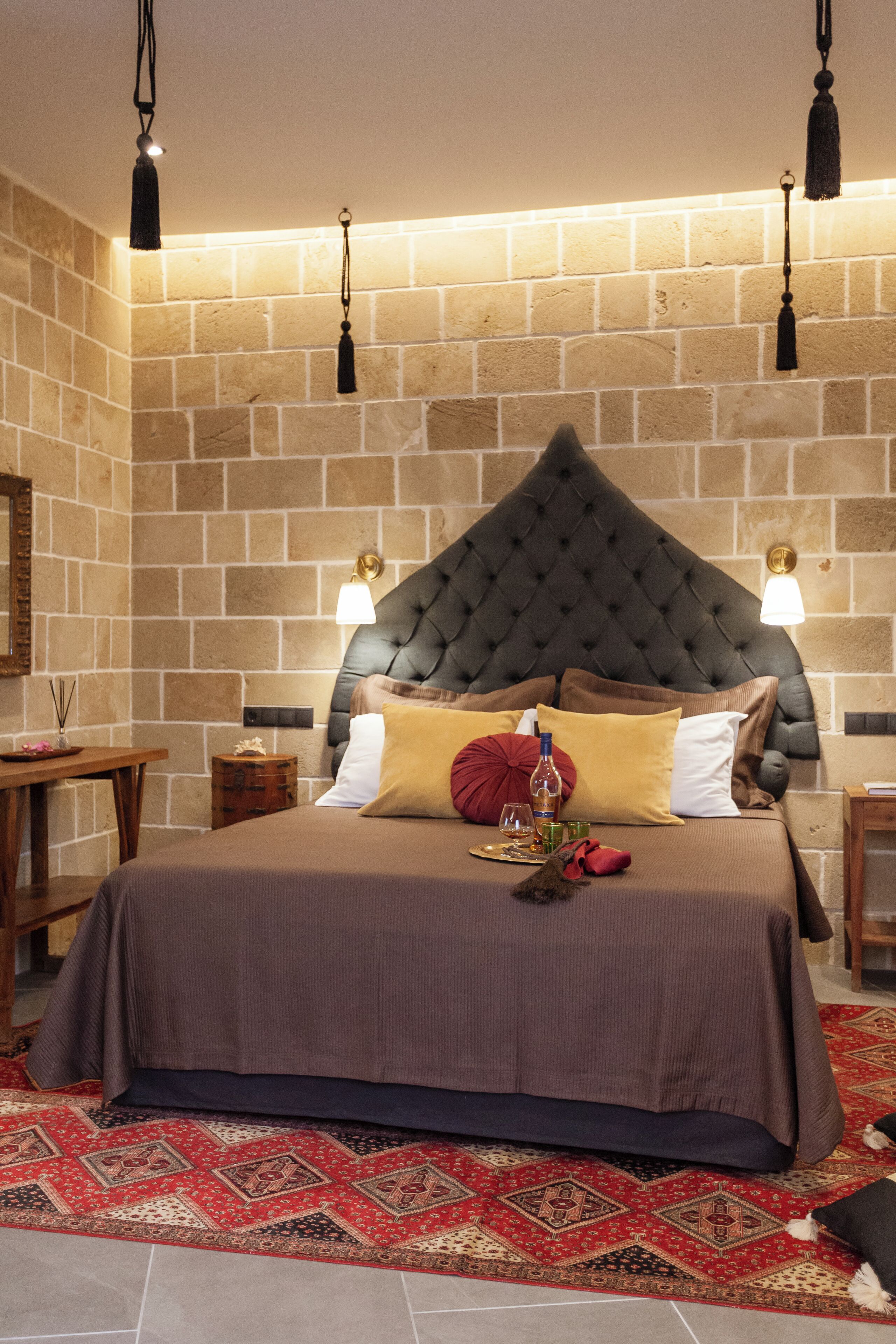 A bedroom with a stone brick wall, a tufted headboard, a patterned rug, and a bedside table with a lamp.