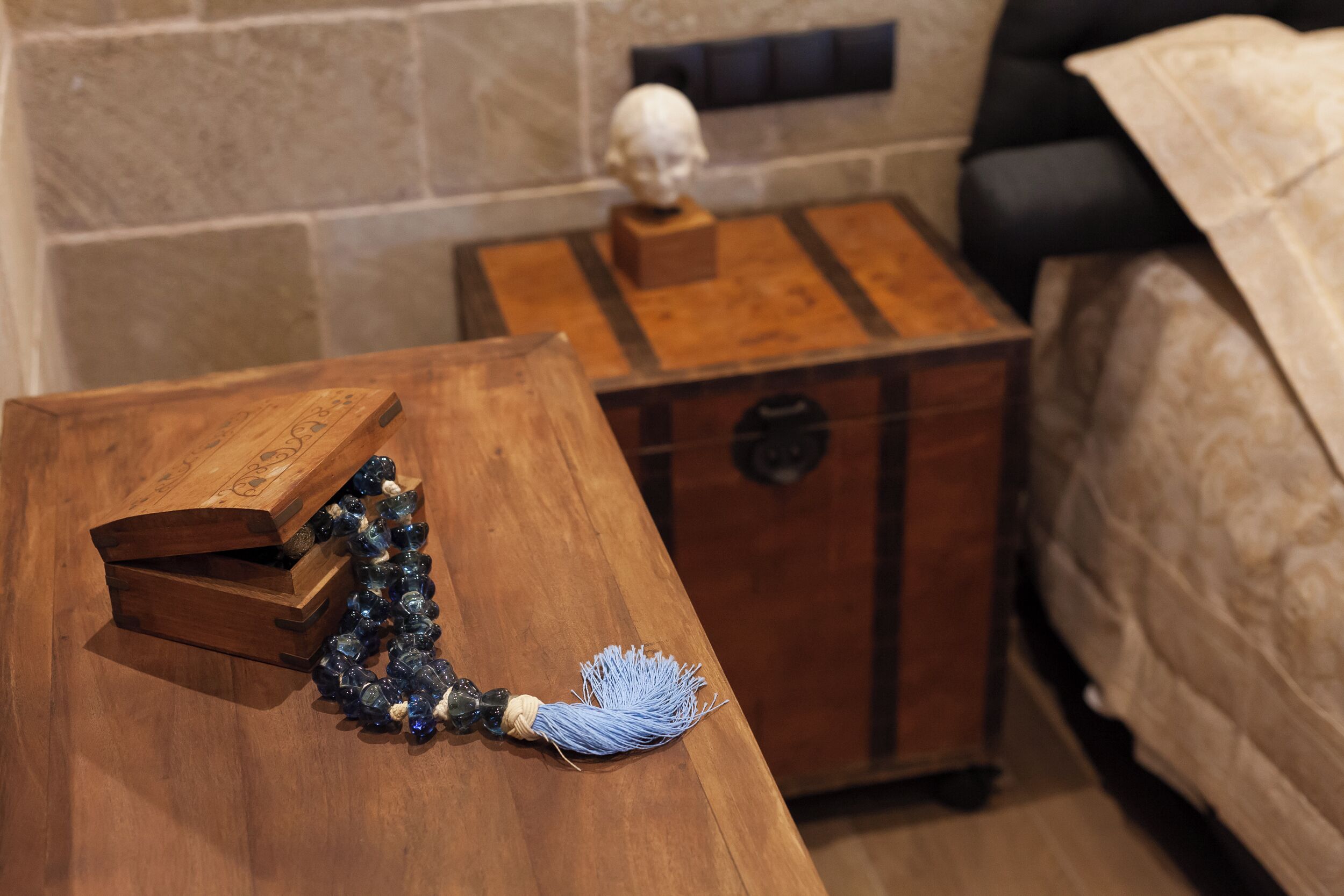 A wooden chest with a blue tassel, a small wooden box, and a bust on a wooden stand in a room with a bed and a stone wall.
