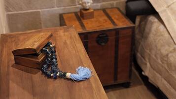 A wooden chest with a blue tassel, a small wooden box, and a bust on a wooden stand in a room with a bed and a stone wall.