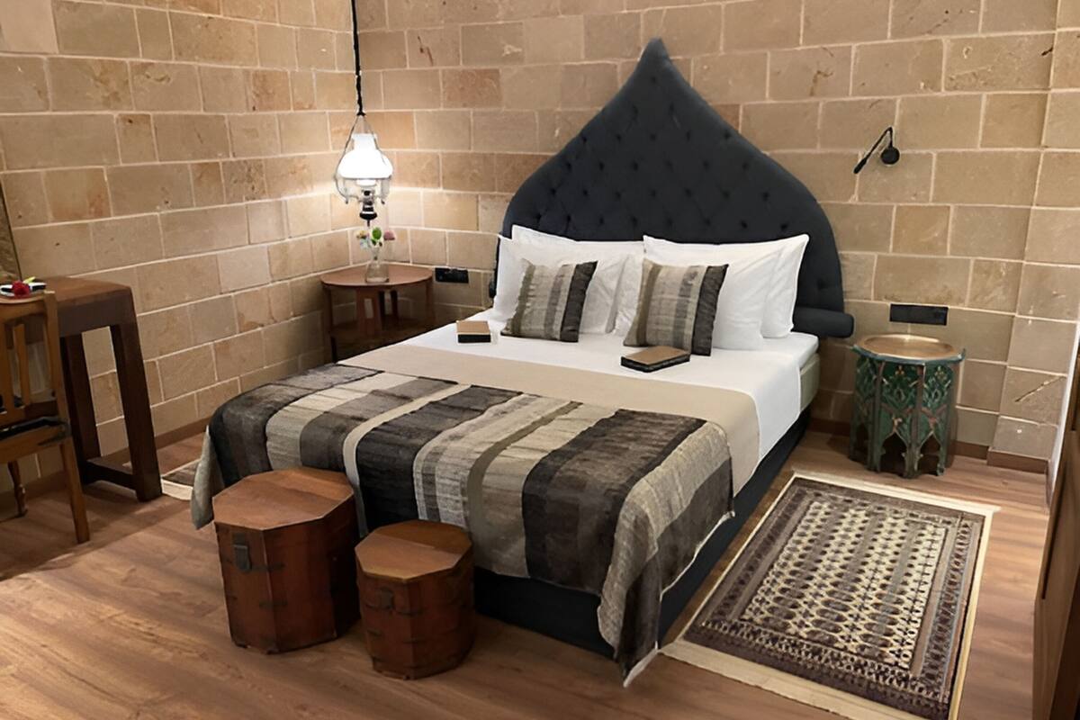 Un dormitorio con cama, dos taburetes de madera, una mesita de noche, una lámpara y una pequeña mesa.