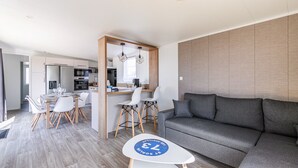 A modern living room with a grey sofa, a dining area with white chairs, and a kitchen in the background. - Holiday at 6 in a résidence with heated pool (Bretignolles-sur-Mer)