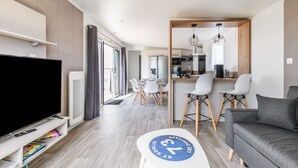 A modern living room with a flat-screen TV, a sofa, a dining area with a table and chairs, and a kitchen in the background. - Holiday at 6 in a résidence with heated pool (Bretignolles-sur-Mer)