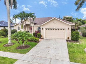 A residential house with a garage, a driveway, and a well-maintained lawn with palm trees. - Pet-Friendly Home | Pool, Spa & Steps to Beach (Marco Island)