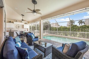 A covered outdoor area with a pool, wicker furniture, and a view of palm trees.