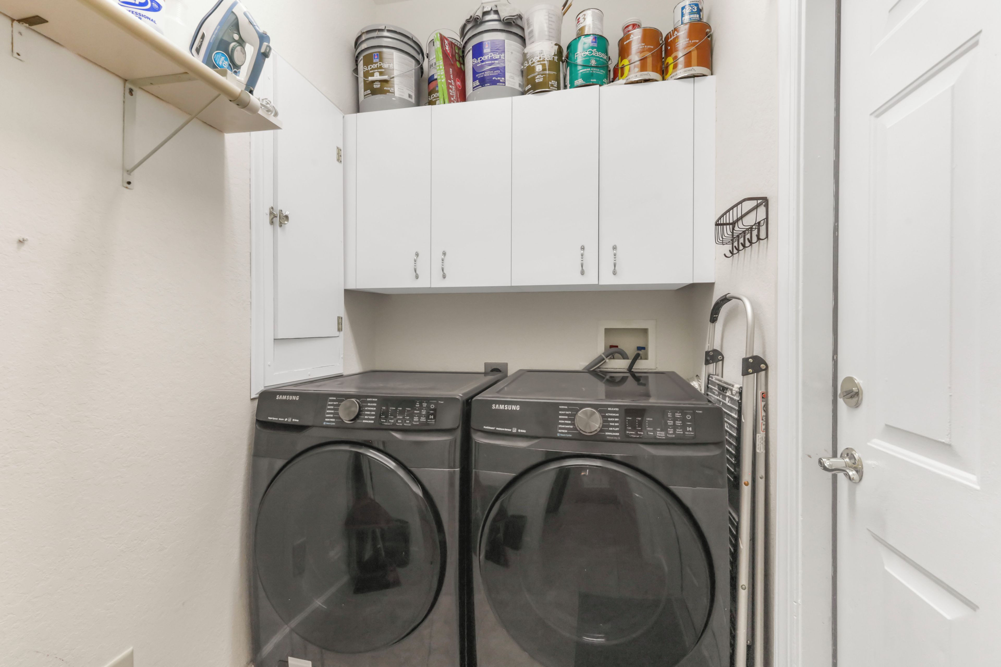 A laundry room with two Samsung washing machines, a white door, and a shelf with various paint cans.