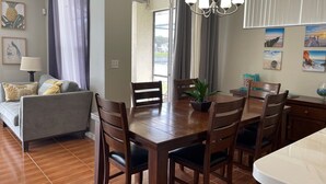 Un área de comedor con una mesa de madera, sillas y una planta en maceta. Hay un sofá con cojines y una lámpara al fondo.