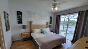 Un dormitorio con cama, mesitas de noche, ventilador de techo, ventana con cortinas y vista a una piscina.