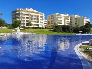 Ein Schwimmbecken mit gefliestem Sonnendeck, umgeben von einer Rasenfläche und einem mehrstöckigen Gebäude mit Balkonen.