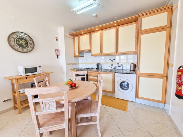 A compact kitchen with wooden cabinets, a microwave, a washing machine, and a dining area with a table and chairs.