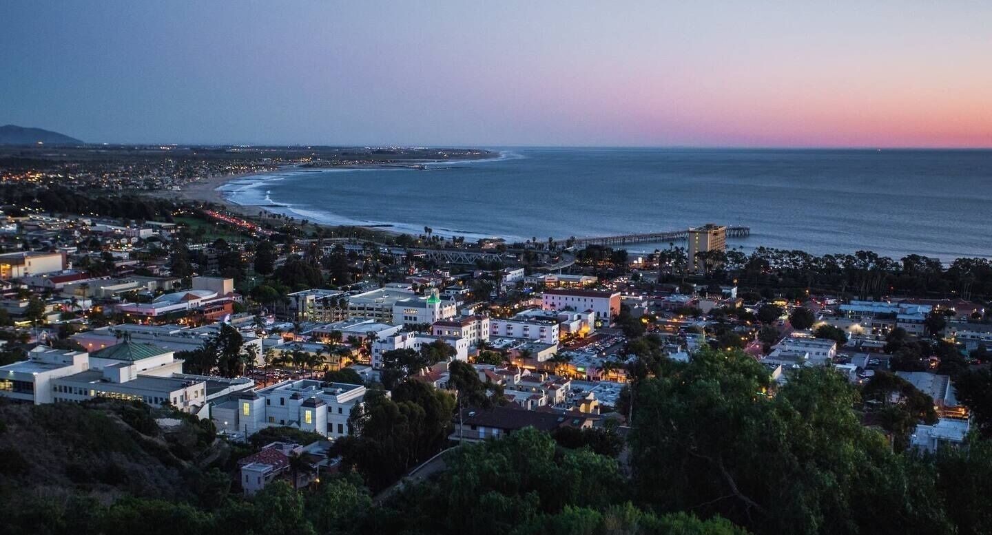 A coastal cityscape at dusk with a prominent pier extending into the ocean.