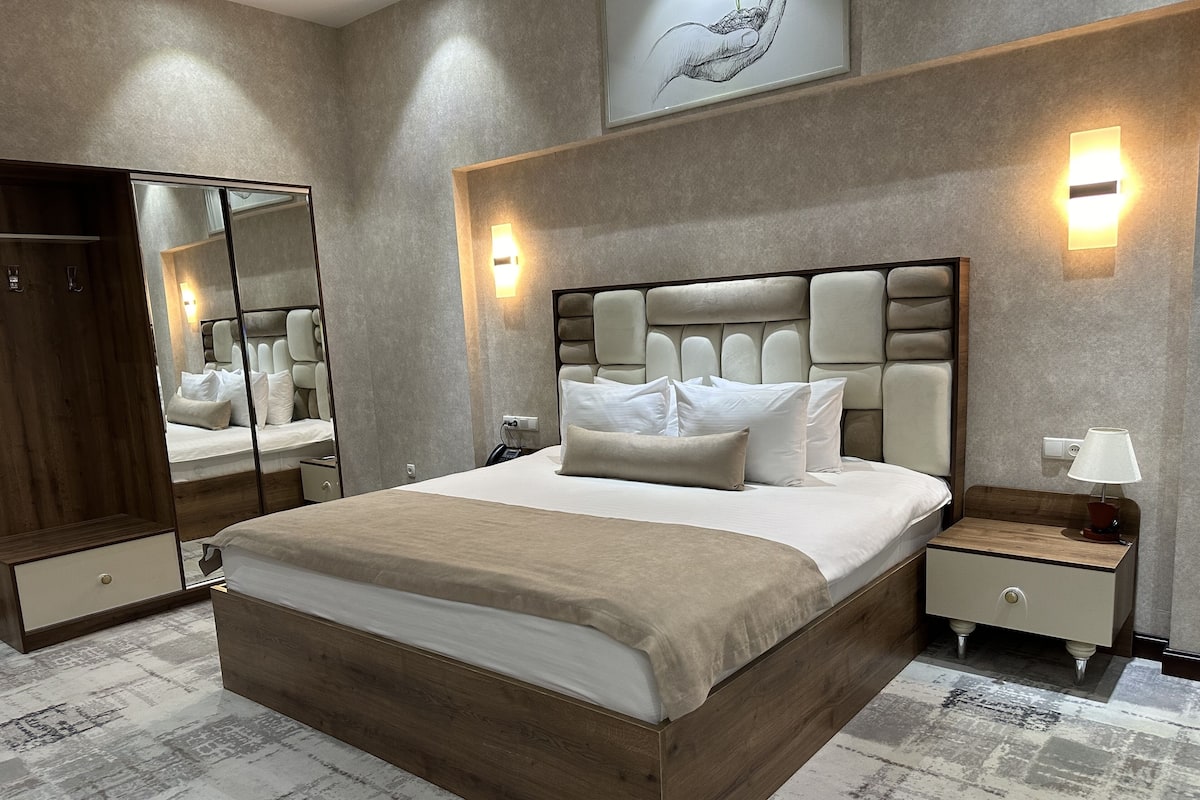 A modern bedroom with a large bed, a wooden headboard, bedside tables, a nightstand, a mirror, and a wall-mounted light fixture.
