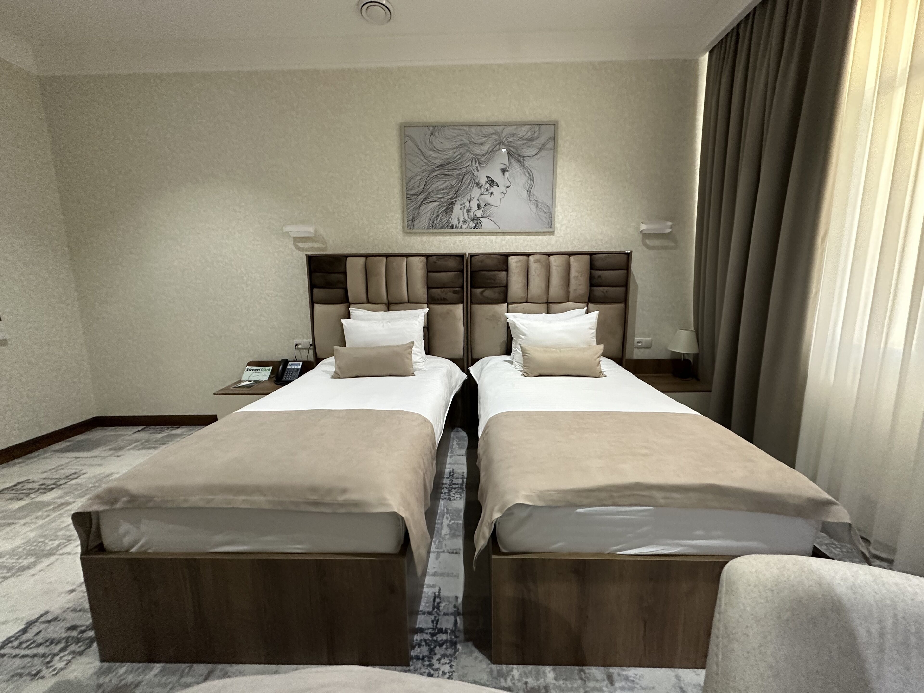 A hotel room with two beds, a headboard with a floral design, a nightstand with a phone, and a painting on the wall.