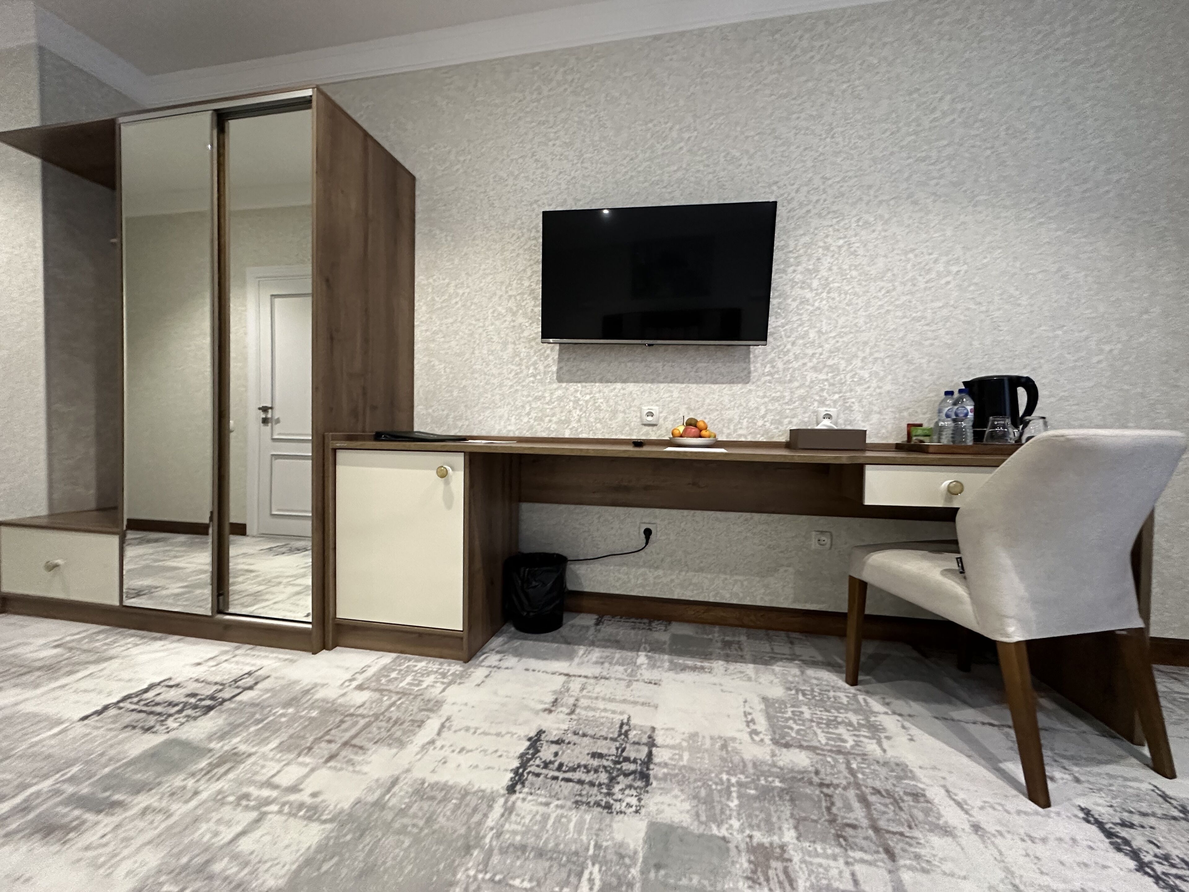Una habitación de hotel moderna con un escritorio de madera, televisión, espejo y un pequeño rincón de descanso.