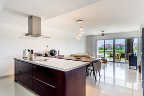 A modern kitchen with a central island, a dining area with a sofa and chairs, and a view of a residential building through large windows.