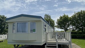 A mobile home with a porch and a bench, set on a gravel area with grass and trees in the background.