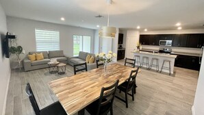 A spacious living area with a wooden dining table, grey sofas, and a kitchen with dark cabinets and a stainless steel refrigerator. - Treasure Coast Retreat 3 bedroom duplex home (PORT ST LUCIE)