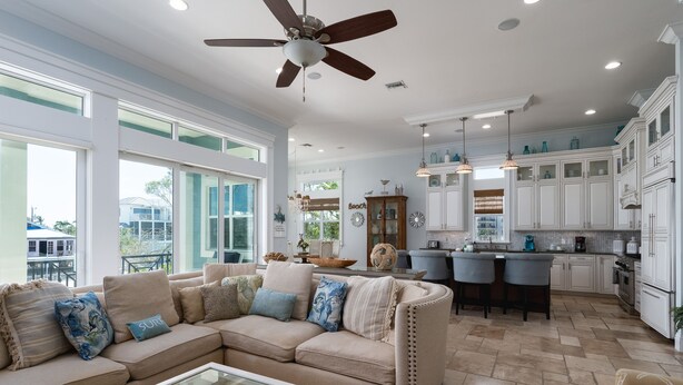 A spacious living room with a large beige sectional sofa, a glass coffee table, and a ceiling fan. Adjacent is a kitchen with white cabinets, a bar area, and pendant lighting.