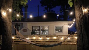 A hammock under string lights, a white building with windows, and trees.