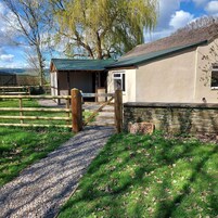 Un bâtiment rustique, doté de fondations en pierre et d’un toit végétalisé, entouré d’une clôture en bois et d’un chemin de gravier menant à un portail en bois.