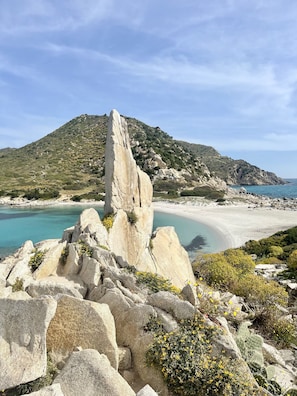 Een kustlandschap met een opvallende rotsformatie, een zandstrand en een glooiend achtergrondlandschap.