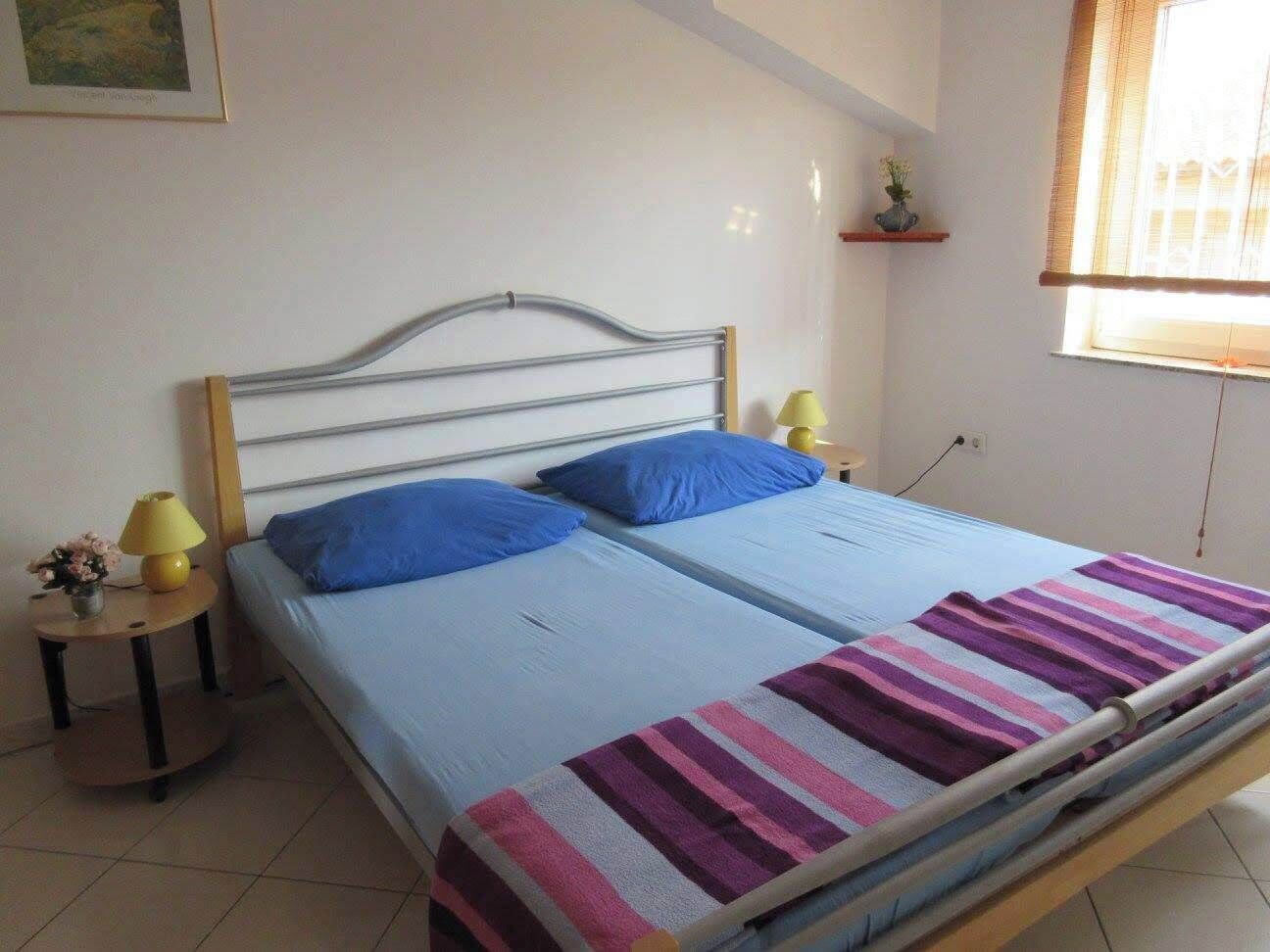 Ein Doppelbett mit gestreifter Decke, zwei Kissen, ein Nachttisch mit Lampe und ein kleines Regal mit einer Pflanze.
