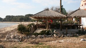Un restaurant situé en bord de mer, doté de parasols en chaume et de tables en bois, offrant une vue sur une plage de sable et l’océan.