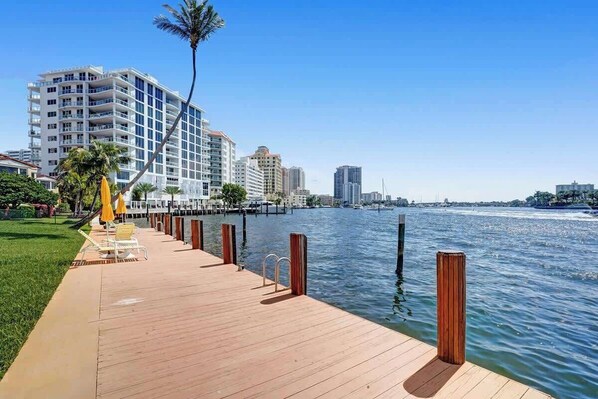 A waterfront promenade with a wooden deck, palm trees, and a view of high-rise buildings across the water.