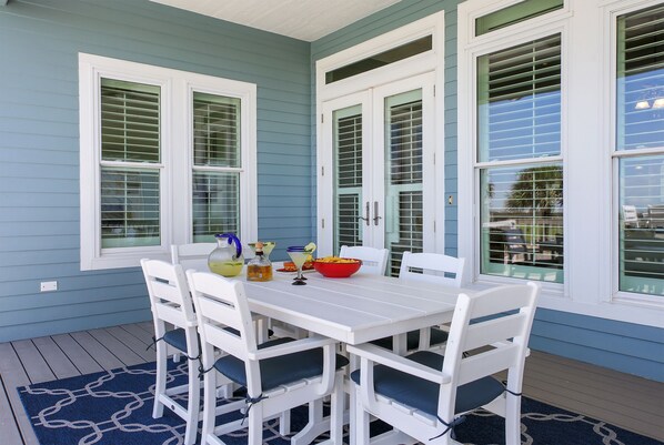 Un coin repas avec une table et des chaises blanches, un tapis à motifs bleus et une fenêtre avec des volets blancs.