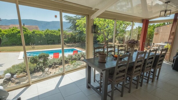 A dining area with a table and chairs, large windows with a view of a swimming pool and greenery, and a ceiling fan. - Villa with pool at the foot of the Luberon (Maubec)
