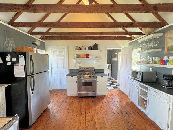A kitchen with wooden beams, a refrigerator, stove, microwave, and various kitchen items on shelves. - Bunting House - Waterfront - Close to town . Pet friendly. (Chincoteague)