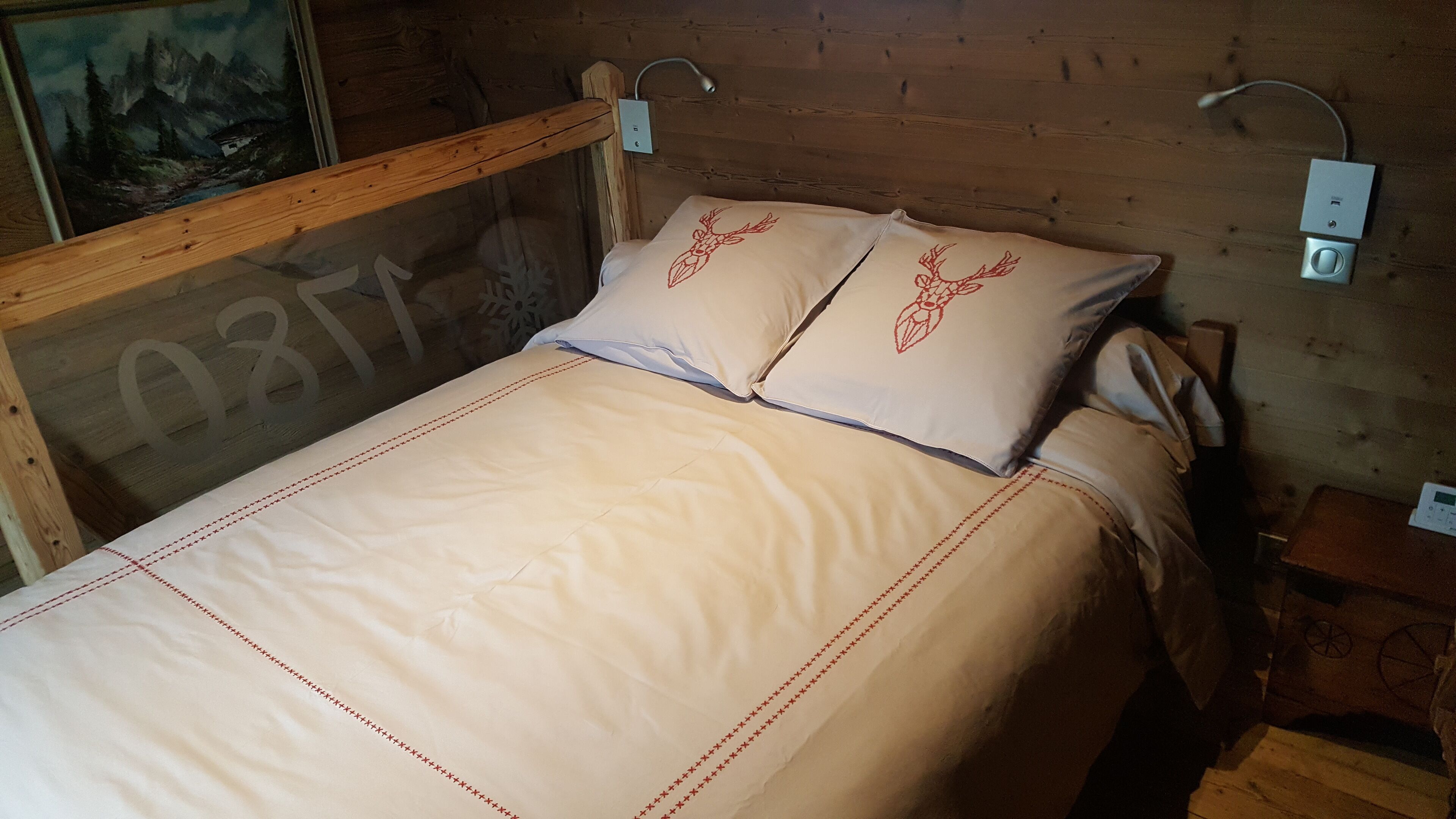Ein Bett mit Holz-Kopfteil, zwei mit Hirsch-Motiv versehene Kissen und ein Nachttisch mit Lampe.