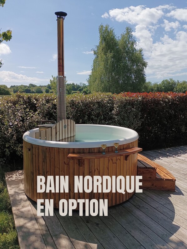 Un jacuzzi de madera con chimenea, rodeado por una terraza de madera y setos, con un cielo despejado al fondo.