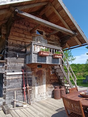 Una cabaña rústica de madera con un porche, una escalera que lleva a una zona de altillo, y un pequeño espacio al aire libre con mesa y sillas.