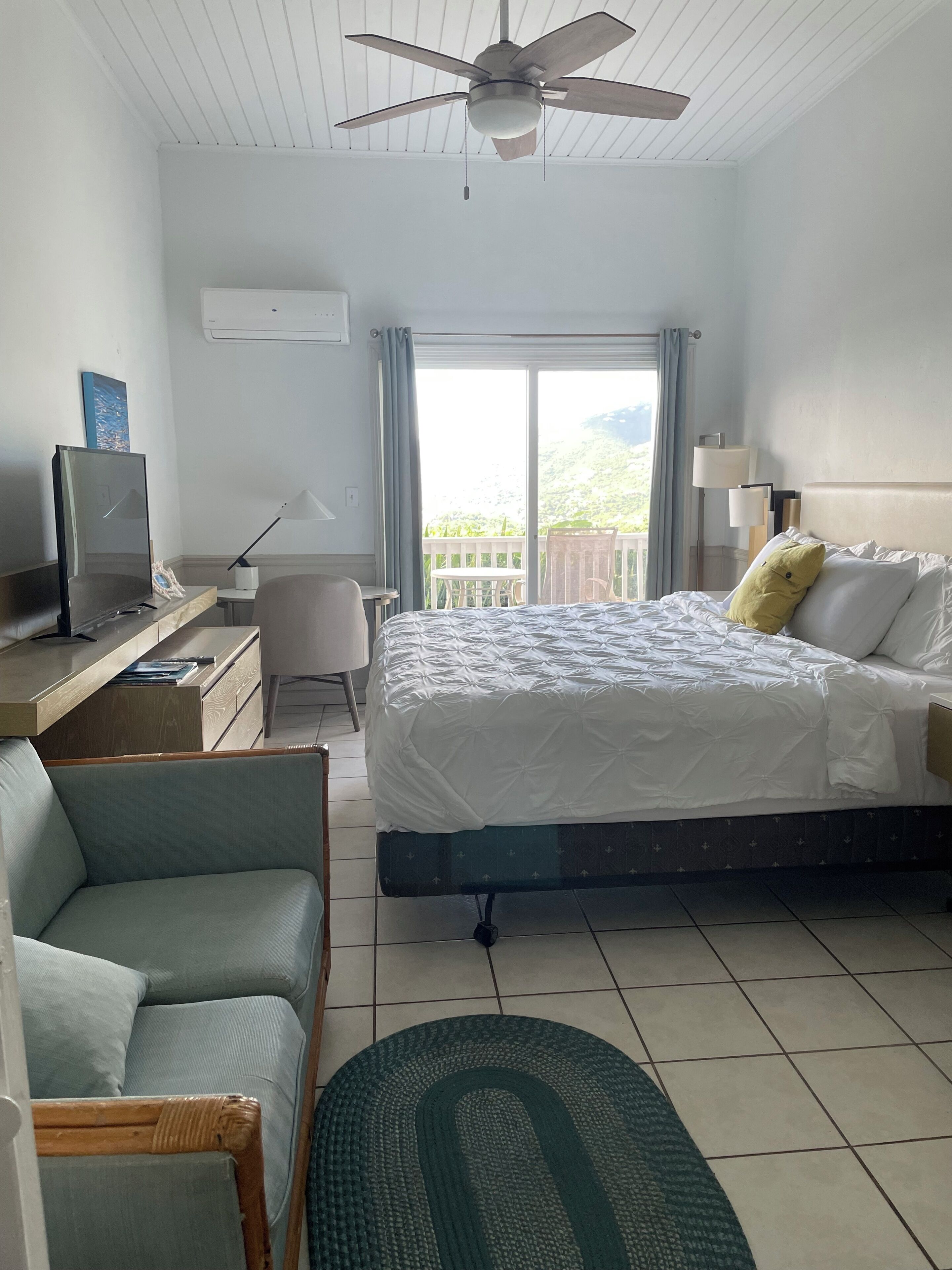 A hotel room with a bed, a desk with a computer, a chair, a TV, a ceiling fan, and a view of the outdoors.