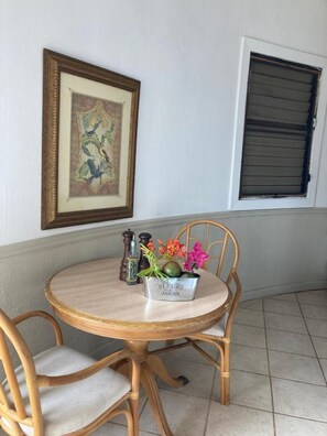 A round wooden table with a metal bowl labeled "FLEURS JARDIN" containing flowers and a green fruit, flanked by two wooden chairs. A framed artwork hangs on the wall above the table. - Boundless Bliss Hotel (St. Thomas)