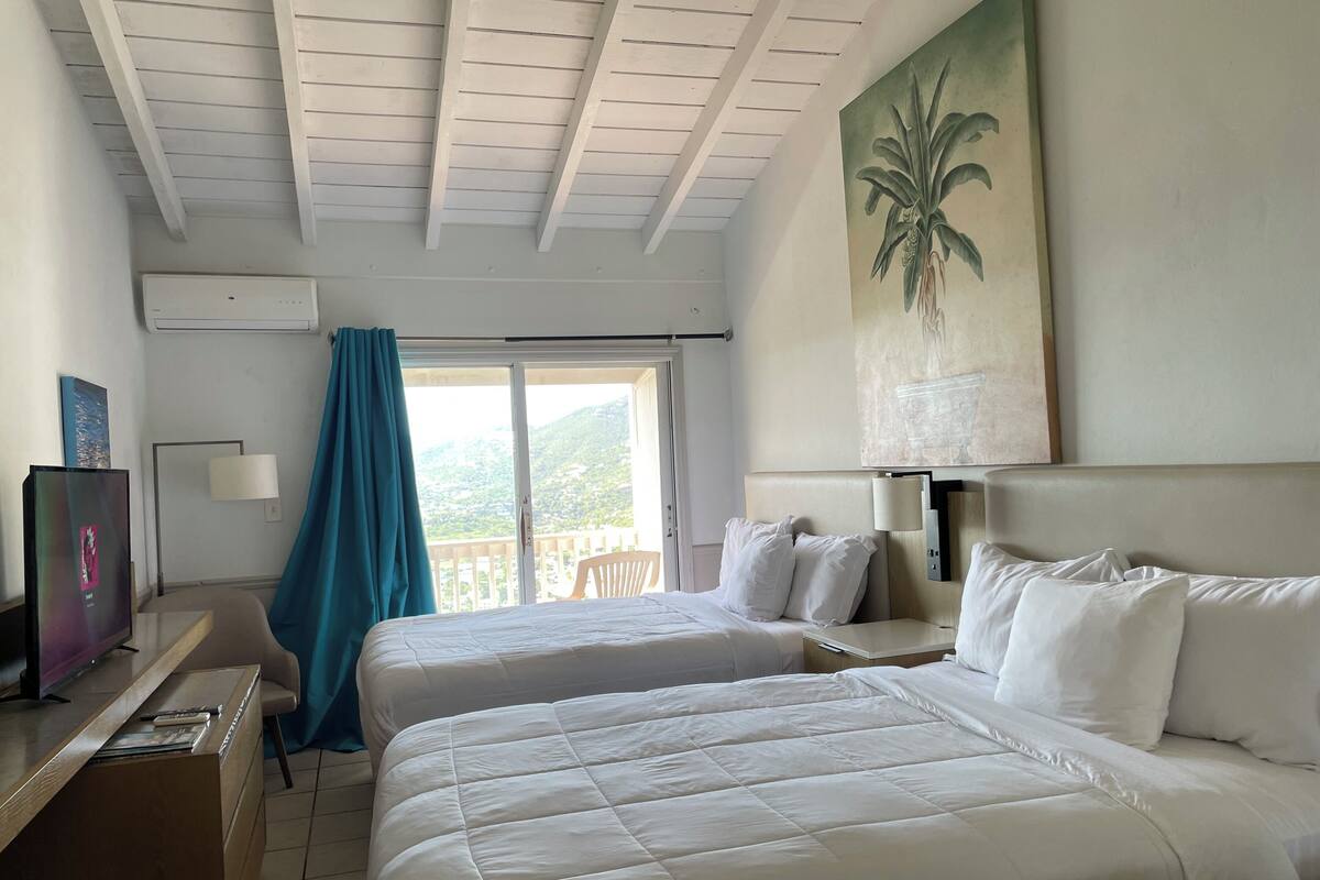 Habitación de hotel con dos camas, un escritorio, un televisor y un balcón con vistas a las montañas.