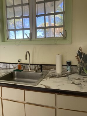 A kitchen with a sink, a window with a view of trees, and a countertop with a soap dispenser and a salt container. - Boundless Bliss Hotel (St. Thomas)