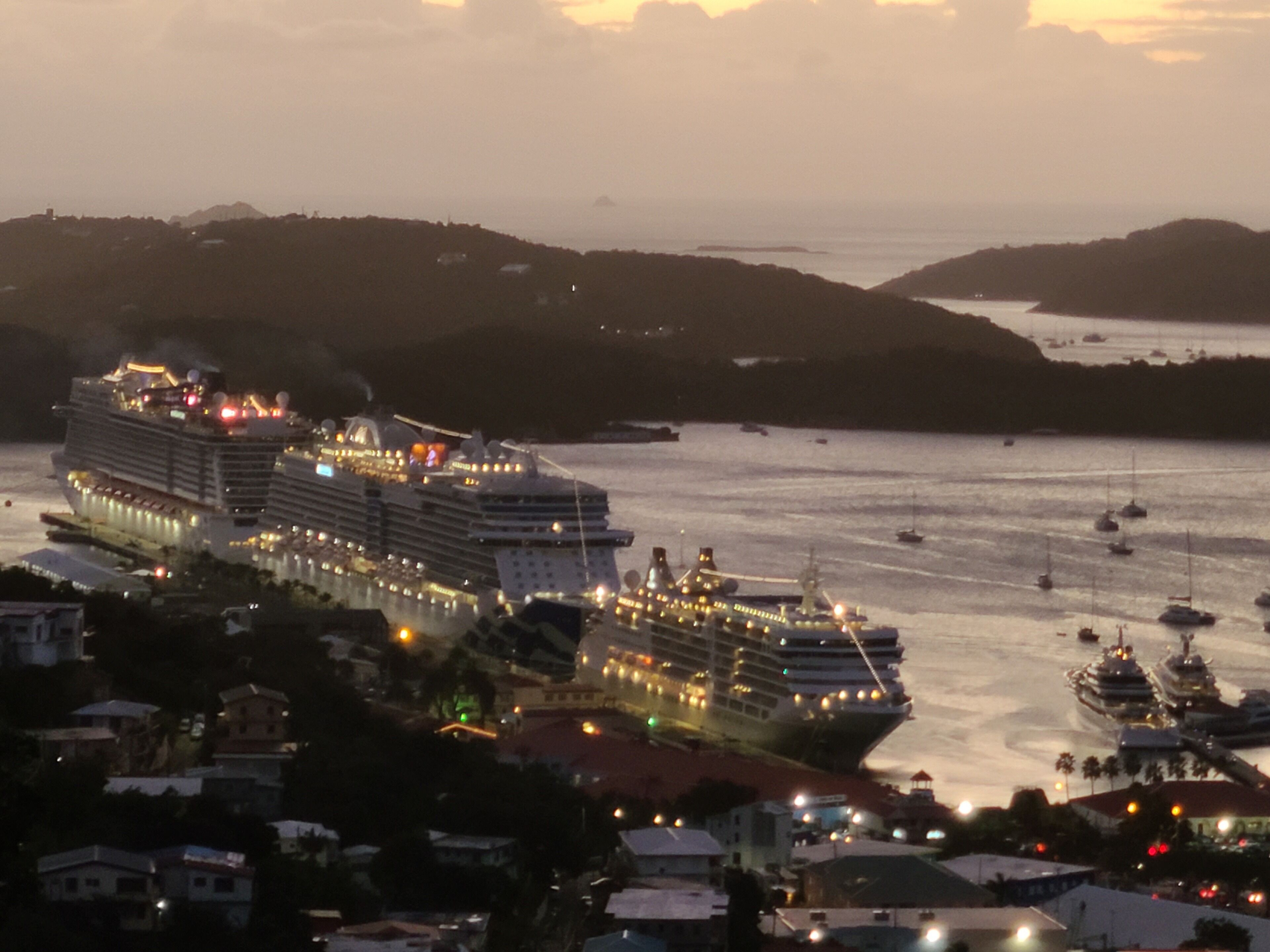 A large cruise ship docked at a port with multiple decks and cabins, surrounded by smaller boats and buildings along the coastline.